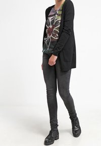Cardigan nero sopra una maglietta grafica con fiori, abbinato a jeans skinny scuri e stivali neri texturizzati. L'outfit presenta una vestibilità rilassata e dettagli discreti.