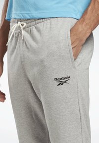 Pantalones de chándal grises hechos de tela suave, con una cintura elástica y un cordón blanco, además de un logo bordado de Reebok en negro en el muslo.