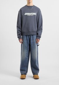Felpa grigia con logo, vestibilità rilassata, polsini a costine, abbinata a jeans blu larghi e calzature beige con suola testurizzata.