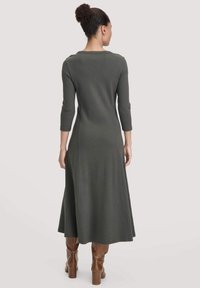 Frau mit lockigem Haar in einem Dutt, die ein langärmeliges, dunkelgrünes Midikleid und braune Absatzstiefel trägt, steht mit dem Rücken zu einem schlichten Hintergrund.