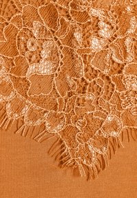Tessuto arancione con dettagli in pizzo intricati sul bordo superiore, caratterizzato da motivi floreali e frange delicate. Texture complessiva liscia.