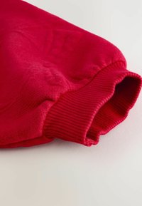 Sweatshirt rouge en tissu doux et texturé. Il possède des poignets côtelés et un col arrondi. Design global lisse sans motifs marquants.
