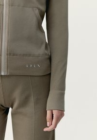 Giacca a zip e pantaloni attillati realizzati in un tessuto a coste dai toni terrosi. Presenta una tasca e dettagli sottili vicino all'orlo e ai polsini.