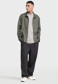 Camisa ligera de color verde oliva con botones sobre una camiseta gris claro, combinada con pantalones de pierna ancha de rayas oscuras y zapatillas blancas.