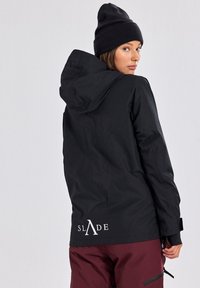 Giacca nera impermeabile con cappuccio, caratterizzata da un logo bianco sulla schiena e polsini elasticizzati. Abbinata a pantaloni borgogna e un berretto nero.