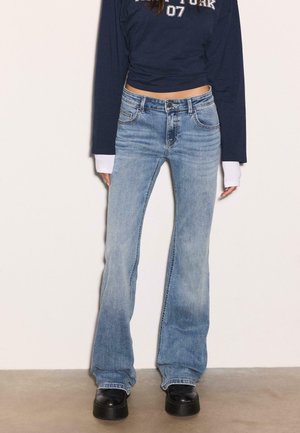 Donna che indossa jeans a zampa d'elefante azzurri chiari, scarpe platform nere e un top corto navy a maniche lunghe con polsini bianchi e testo.