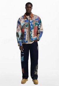 Džínová bunda typu patchwork s mixem živých barev a vzorů, doplněná tmavými kalhotami a béžovou obuví. Hnědý detail límce.