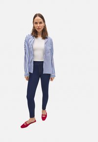 Camisa a rayas azul y blanca, camiseta blanca, leggings azul oscuro y zapatos rojos de slip-on. La camisa tiene un bolsillo y un corte holgado.