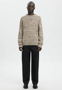 Hellbrauner Strickpullover mit strukturiertem Muster, gerippten Bündchen und Saum, kombiniert mit schwarzen Hosen mit weitem Bein und schwarzen Schuhen.