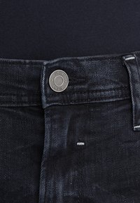 Mörka denimjeans har en metallknapp med "BLEND" präglat, kontrasterande sömmar och subtila slitmärken nära framfickan.