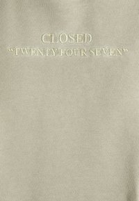 Texte brodé "CLOSED 'TWENTY FOUR SEVEN'" en fil crème sur un fond de tissu beige clair.