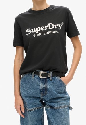 Osoba nosząca czarny T-shirt SuperDry Soho London włożony w wysokie niebieskie dżinsy z ozdobnym czarnym paskiem z srebrną klamrą.