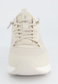 Baskets de sport beiges avec une tige en maille texturée, des lacets plats et une semelle rembourrée. Dispose d'un onglet au talon pour un enfilage facile.