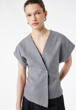 Jeune femme portant un haut gris à manches courtes avec un fermoir argenté, un pantalon noir et des boucles d'oreilles argentées en forme d'anneau sur fond blanc.