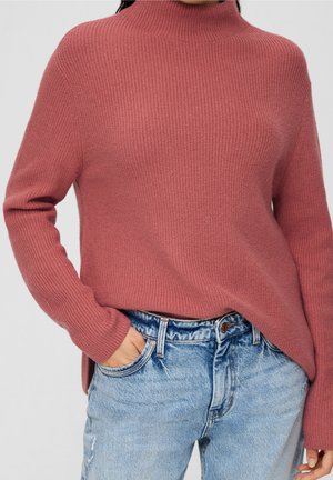 Kobieta ubrana w prążkowany, brudnoróżowy sweter z golfem i jasnoniebieskie jeansy z wysokim stanem, z jedną ręką w kieszeni.