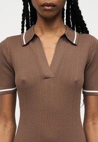 Άτομο που φοράει καφέ κοντομάνικο πόλο με ανάγλυφες ρίγες και λευκή επένδυση στο γιακά και στις άκρες των μανικιών, φαίνεται από τους ώμους μέχρι το μέσο του κορμού.