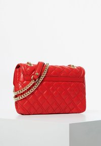 Rød quiltet læderhandbag med en guldkædrestrop. Har en klap-top og en baglomme, kendetegnet ved diamantmønstret syning.
