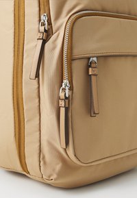 Beiger Nylon-Rucksack mit glatter Textur, ausgestattet mit einem großen Reißverschlussfach und einer Vordertasche. Akzente sind braune Leder-Ziehschlaufen.