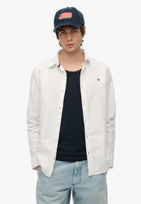 Camisa blanca de botones sobre una camiseta sin mangas azul marino, combinada con jeans de un azul claro. Una gorra azul marino presenta un diseño bordado de la bandera americana.