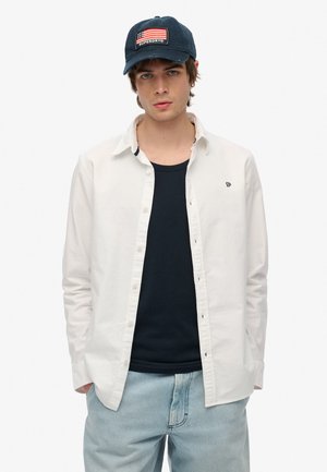 Witte button-up shirt over een marineblauwe tanktop, gecombineerd met lichtblauwe jeans. Een marineblauw pet heeft een geborduurd ontwerp van de Amerikaanse vlag.