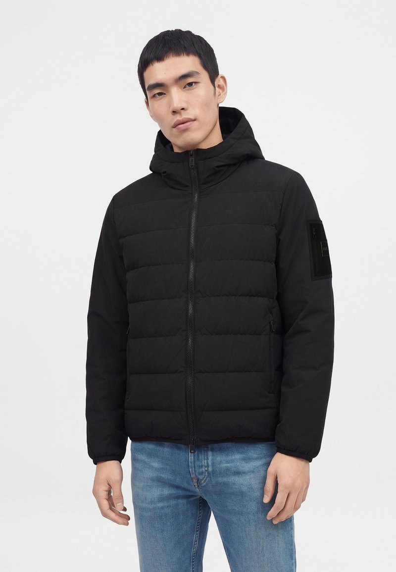 Jeune homme portant une veste matelassée noire zippée à capuche et un jean bleu clair, debout devant un fond clair uni.