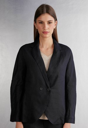 Blazer de lino negro con un diseño estructurado, que presenta un cuello con muescas, cierre de doble botón y mangas largas. Debajo, una blusa de color beige claro.