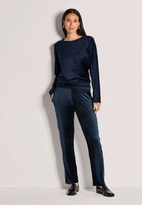 Vrouw in een marineblauwe fluwelen top met lange mouwen en subtiele luipaardprint, met bijpassende marineblauwe fluwelen broek en zwarte instappers, staand tegen een witte muur.