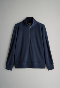 Pull à manches longues en tricot côtelé bleu marine avec un col à demi-fermeture éclair à l'avant, posé à plat sur une surface gris clair.