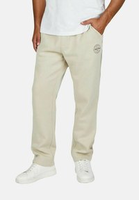 Beige Jogginghose in lockerer Passform, mit zwei Seitentaschen und einem kleinen Logo an der linken Oberschenkel. Getragen mit weißen Turnschuhen.