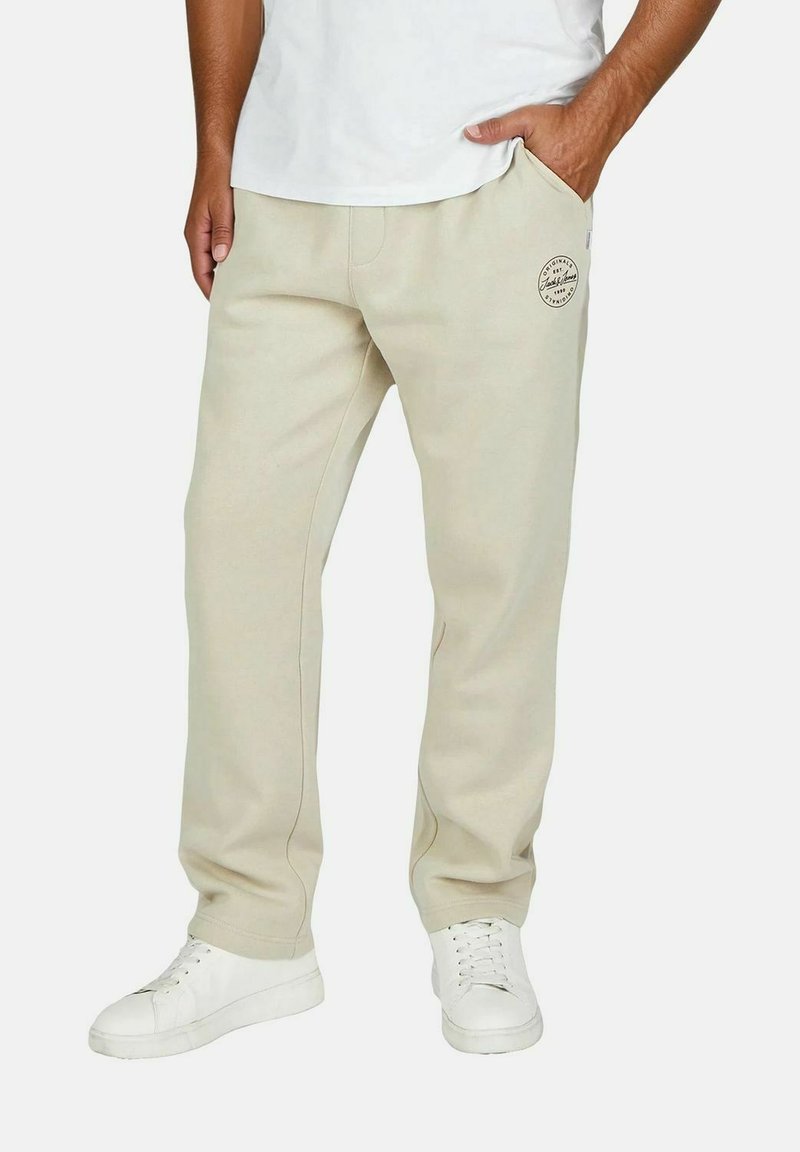 Beige Jogginghose in lockerer Passform, mit zwei Seitentaschen und einem kleinen Logo an der linken Oberschenkel. Getragen mit weißen Turnschuhen.