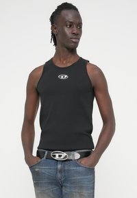 Μαύρη αμάνικη μπλούζα με λογότυπο στο μπροστινό μέρος, συνδυασμένη με μπλε τζιν παντελόνι με εφέ φθοράς και μαύρη ζώνη με ασημένια πόρπη.