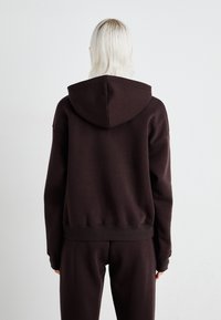 Hoodie castanho feito de tecido macio, com capuz ajustável por cordão e bainha canelada, combinado com calças de fato à correspondência. Design minimalista, sem logótipos visíveis.