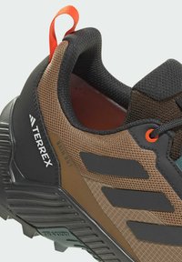 Brown and black athletic shoe with textile upper, rubber sole, and textured patterns. Features orange accents and a pull tab on heel.