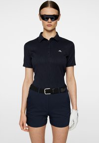Marinblå polo med strukturerat mönster, tre knappar och en logotyp. Kombinerad med matchande marinblå shorts, ett svart nätbälte och en vit golfhandske.