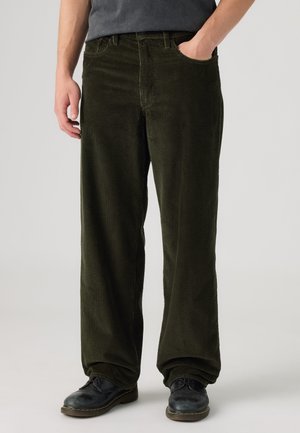 Homme portant un pantalon large en velours côtelé vert foncé, des chaussures en cuir noir et une chemise gris foncé, main dans la poche.