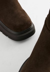 Bottes chevilles en suède marron avec un bout rond, une semelle en caoutchouc noir épaisse et une texture lisse. La surface présente une finition mate douce.