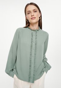 Blusa verde claro con cuello acampanado y cierre de botones en la parte frontal, que presenta un detalle texturizado a lo largo de la tapeta y mangas fruncidas.