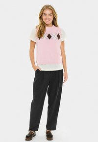 Haut rose clair à manches courtes avec un motif en losanges argyle, associé à un pantalon noir ample et des mocassins marron.