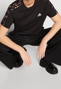 Černé tričko s krátkým rukávem, které má na ramenou tři vertikální pruhy v kostkovaném vzoru a vyšitým logem Adidas na hrudi.