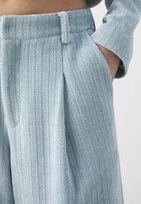 Personne portant un pantalon en maille texturée bleu clair et un haut à manches longues assorti, main reposant négligemment dans la poche.