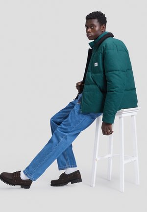 Ung mand i grøn dynejakke, blå jeans, hvide sokker og brune sko sidder sidelæns på en hvid skammel mod en grå baggrund.