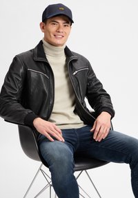 Zwarte leren jas met zilveren ritsen, beige coltrui en donkerblauwe jeans. Het model draagt een blauwe pet met een gele logo, zittend op een stoel.