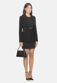 Blazer nero su misura con dettagli in oro, abbinato a un vestito nero aderente e tacchi con cinturino alla caviglia. Tiene una borsa nera con logo dorato.