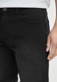 Jean en denim noir avec une coupe slim, présentant des passants de ceinture, une poche avant et un ourlet retourné. Texture lisse avec un minimum de détails de couture.