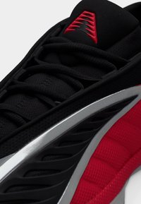 Zapatilla atlética negra con acentos rojos, tejido texturizado, detalles plateados y un diseño dinámico que presenta cordones prominentes y patrones de cuadrícula.