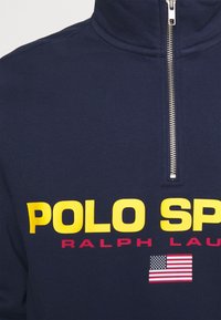 Navyblaues Zip-Hoodie aus Baumwolle, mit großem gelbem "POLO SPORT" Schriftzug, rotem "RALPH LAUREN" Schriftzug und einer kleinen amerikanischen Flagge.