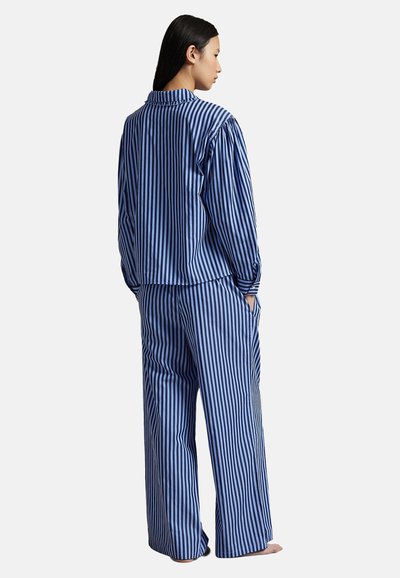 Femme debout pieds nus, portant une chemise ample à manches longues à rayures verticales bleues et noires assortie à un pantalon large, les mains dans les poches.