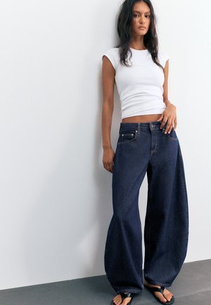 Donna che indossa un top bianco senza maniche e jeans scuri a gamba larga, in piedi a piedi nudi contro un muro bianco e pavimento grigio.