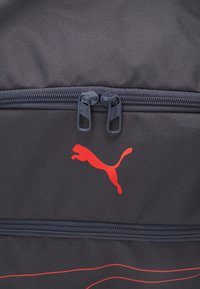 Crni platneni ruksak s crvenim detaljima. Ima istaknuti crveni Puma logotip i sive ručke za zatvaranje. Glatka tekstura s horizontalnim uzorcima.