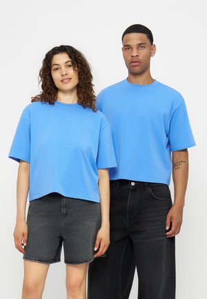 Ung mand og kvinde står side om side iført matchende lyseblå cropped t-shirts og sorte jeansbukser mod en ensfarvet hvid baggrund.